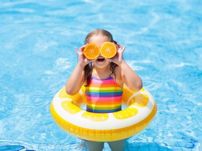 Child in swimming pool. Kid eating orange. Little girl in swimming pool with inflatable toy ring eating orange. Kids swim on summer vacation. Tropical fruit and healthy snack. Swim aids for child. Kid on colorful float. Beach and water fun.